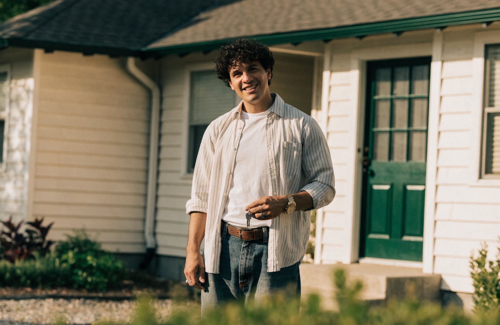 A landlord holding keys, smiling in front of a well-kept rental home on a sunny residential street